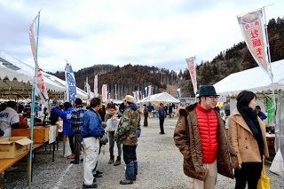背景には木々が生い茂る山並みが広がり曇り空の中、白いテントの間には多くの人々が行き交り、買い物客が立ち止まって商品を見たり店員と話したりしている様子を写した写真