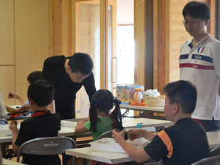 長机に座ってロボット作りをしている子供たちを横に立って見ている講師の猪狩憲一さんと、黒いシャツを着た男性が女の子に教えている様子を写した写真