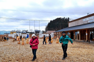 女性はピンクのダウンジャケットと白いニット帽を着用し隣を歩く男性はエメラルドグリーンの防寒着と黒い帽子を着けた男女がカメラに向かって歩いてくる仮設商店街の一角にある屋外広場を写した写真