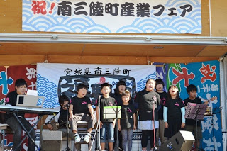 「南三陸町産業フェア」と書かれた横断幕が掲げられたステージで、お揃いの黒いシャツを着た子どもたちがマイクの前に並んで合唱している写真