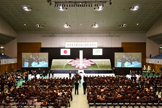 広々とした室内会場で、多くの参列者が椅子に座って前を向き、正面には大きな花の祭壇と日本国旗、二つのスクリーンに映像が映し出されている追悼式の写真