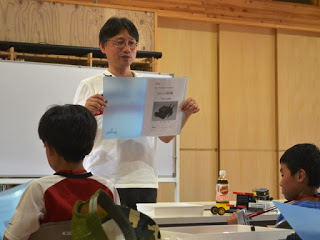 教室で猪狩憲一さんが資料を掲げながら子どもたちに説明している様子の写真
