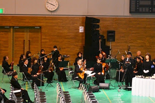 追悼式の会場で弦楽器や管楽器を持った演奏者たちがオーケストラ演奏をしている様子の写真