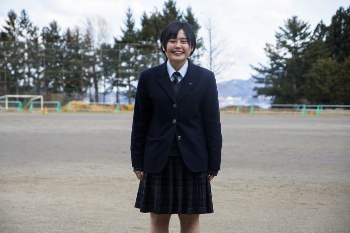 グラウンドの中央でブレザーの制服を着た女子高校生の三浦千裕さんが笑顔で立っている写真