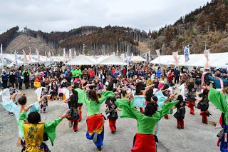 背景には山々が広がっている仮設テントが並ぶ広場の中央で、カラフルな赤、青、黄など鮮やかな色合いの衣装を着た複数の踊り手（主に若者や子ども）が、観客に向かって元気よく踊っている様子を写した写真