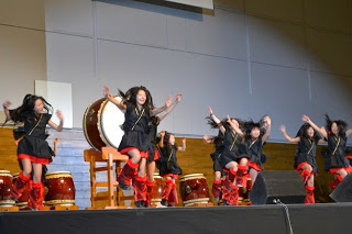 太鼓を並べた舞台の上でお揃いの衣装を着た人たちが両手を上にあげ踊っている写真