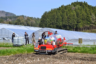2人がカメラを構えて撮影している中、イラスト付きの液肥専用散布車が畑の中を進んでいる様子を写した写真