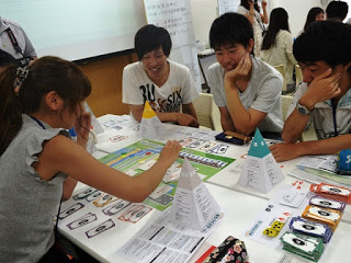 加者たちがテーブルを囲んで「みんなのまちづくりゲーム」を楽しんでおり、女性がカードを出して周囲が笑顔で見守っているボードゲーム中の写真