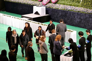 追悼式の様子で、中央の白いテーブルには花が飾られ、壇上には黒い布で覆われた台があり、人々が列を作って献花をしている場面の写真