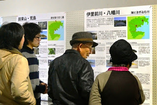 数人の人々が「伊里前川・八幡川」や「田束山・馬島」といったタイトルの展示パネルを見ている様子を写した写真