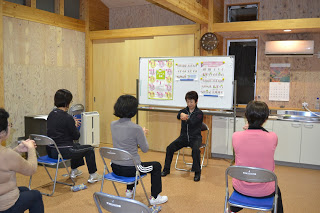 木の壁の室内で、椅子に座った数人の女性がインストラクターの動きに合わせて軽い運動をしている写真