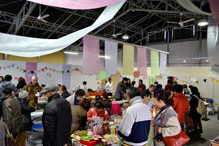 天井から布が飾られた広い屋内会場で、多くの人々が手づくり品のブースを見て回ったり買い物を楽しんだりしている様子が写っている写真