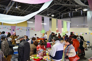 天井からは淡い色（ピンク、黄、白、緑など）の布や飾りが垂れ下がっている会場内で、多くの人々が商品を見たり、店員と会話したりしている様子を写した写真