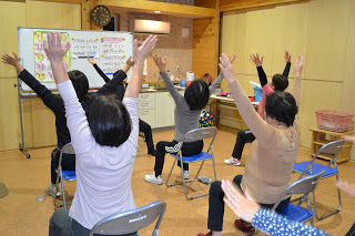 参加者が椅子に座ったまま両手を大きく上に広げて、全員で体操をしている写真