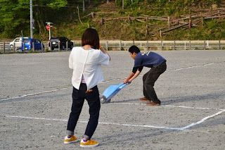 屋外のグラウンドで、背中を曲げて白線を引いている男性の様子を、女性が撮影している写真