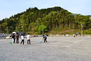 木々が茂った丘の手前にある野外の広いグラウンドで、白線を引いている男性を数人の参加者が見ている写真