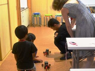 子供たちが床に置いた自走ロボット「ペッパーくん」で遊んでいて、大人の女性と男性がその様子を見守っている写真