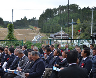 野外で開かれた式典に整列して座り、配布された資料を手にしながら話を聞いている多くのスーツ姿の出席者を写した写真