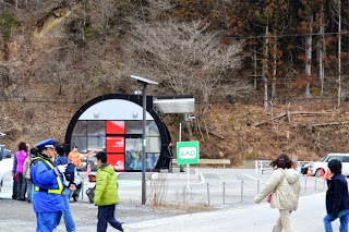 背景には木々の生えた山肌があり、落葉した木々が多く中央奥には、半円形の黒いフレームを持つ小さな建物があり、手前の舗装された道路や歩道の左側には制服を着た係員と、その近くを歩く子どもや大人を写した写真