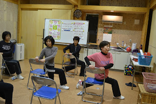 椅子に座った状態で体をひねるような運動を女性たちが行っており、インストラクターも一緒に動いている写真