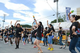 黒いTシャツとショートパンツ姿の女性たちが、カラフルな小道具を振りながら楽しそうに踊りを披露している写真