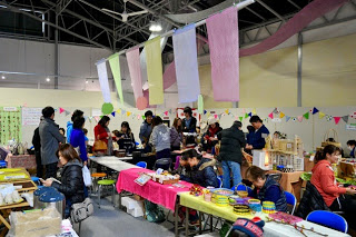 天井から色とりどりの布が吊るされた屋内会場で、多くの人々が手作り品の販売ブースを見て回ったり買い物を楽しんだりしており、テーブルの裏側では出店者たちが商品を並べたり接客をしたりしている様子を写した写真