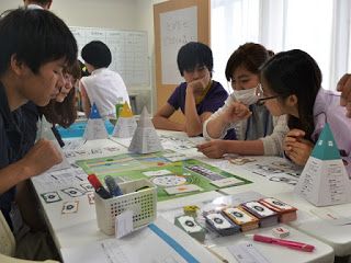 数人がテーブルを囲んでボードゲームに取り組み、カードや資料を見ながらゲームを楽しんでいる様子の写真