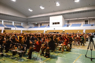 広い体育館内で観客の人たちがパイプ椅子に座っている会場内を写した写真