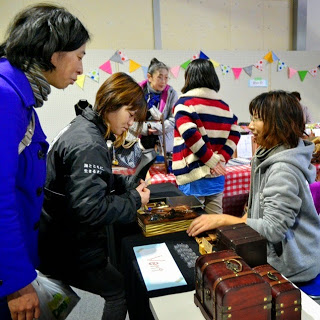 テーブルに並んだ木製の小箱を前に、グレーの服を着た販売者が笑顔で説明をしており、それに対して黒い服の女性と青いコートの女性が話を聞いている、にぎやかな室内会場の一角を写した写真