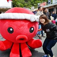 白い鉢巻きを巻いた大きな赤いタコのマスコットの横で、笑顔の女性が少し腰をかがめて立っている写真