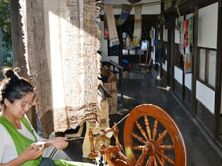 女性が木製の糸車を使って糸を紡いでおり、背景には手織りのカラフルな布が吊るされている古民家の廊下が映っている写真