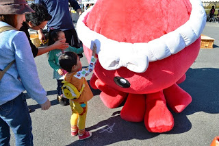 大人たちが見守るなか、赤いタコのオクトパス君の着ぐるみに小さな子どもが手を伸ばして触れている写真