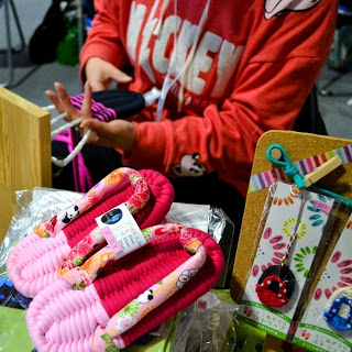 ミッキーのパーカーを着た人がカラフルな布を手に編み物のような作業をしており、手前のテーブルにはピンク色の手作り布ぞうりや花柄の飾りが並べられている様子を写した写真