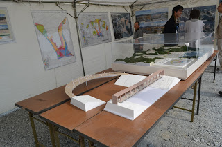 壁に複数の図面や写真が掲示されたテントの中で、テーブルの上に曲線状の中橋等の模型やガラスケースに収められた景観模型が展示されており、男性スタッフが見学者に説明を行っている様子の写真