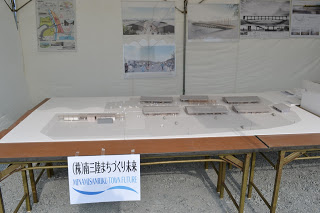 プロジェクトに関する写真が複数貼られた壁の下に設置された「株式会社南三陸まちづくり未来」と書かれた張り紙が掲げられているテーブルの上に、精巧な建築模型が展示されている写真