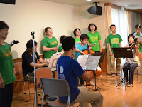 椅子に座って楽器を演奏している4人の後ろに黄緑色のTシャツを着た合唱団が並び、歌を歌っている写真