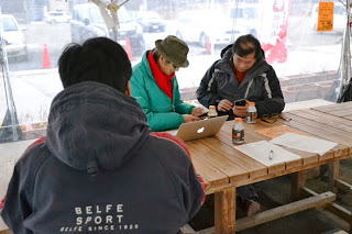 木製の机を囲んで、奥に並んで座る男性2人がノートパソコンを前に置き携帯電話を操作し、手前にもう1人の男性が背中を向けて座っている様子を写した写真