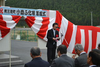 赤と白の幕を背景に、壇上でマイクを使ってスピーチをしている男性と、それを正面から座って聞いている出席者たちを写した写真