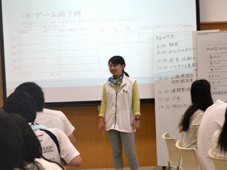 白いベストを着た進行役の女性がスクリーンの前で話しており、参加者が座って話を聞いている中、ホワイトボードにスケジュールやルールが書かれている教室内の写真