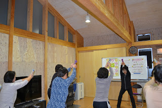 数人の女性がインストラクターの真似をしながら、両手に小さなダンベルを持って天井に向かって腕を伸ばしている写真