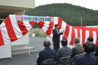 屋外の壇上でスピーチ原稿を手にして話している別のスーツ姿の男性が、出席者に向かって話している赤と白の幕の前の写真