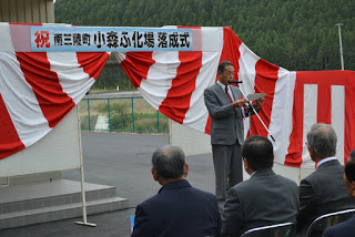 赤いネクタイを締めた来賓の男性が、赤と白の幕の前で紙を読みながらスピーチをしている姿と、それを聞く参加者たちを写した写真