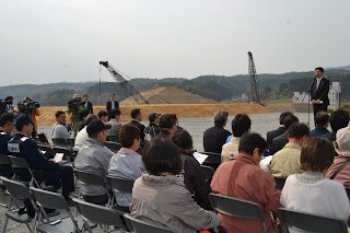 背景には建設用のクレーンや機材が見られる屋外で、たくさんの出席者が広い敷地内に設置された椅子に座り、檀上のマイクの前に立つ男性の話を聞いている見学会の様子の写真