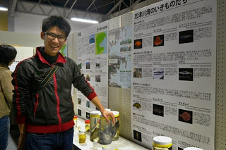 展示会のブースで男性が瓶入りの標本を指し、背景に日本語の説明パネルがある様子を写した写真