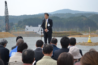 背景には建設用のクレーンや機材が見られる屋外で、設置された椅子にたくさんの出席者が座っており、スーツ姿の男性がマイクの前に立って話している様子を写した写真