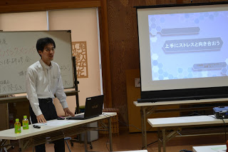 スーツ姿の男性講師がプロジェクターとパソコンを使って、「上手にストレスと向き合おう」と書かれたスライドを示している様子の写真