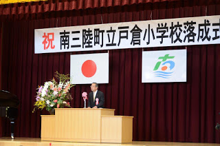 紅白の幕と大きな花が飾られた舞台で、スーツ姿の男性が演台に立ち、落成式のあいさつをしている壇上の写真