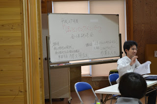 ホワイトボードに「平成27年度 住民いきいきサロン 会話研修会」と書かれ、講師が紙を手に説明している様子の写真