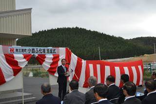 山と空を背景にした屋外式典で、スーツを着た男性がスピーチをし、複数の出席者が真剣に話を聞いている赤と白の幕の前の写真