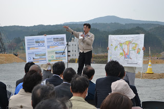 背景には山々や建設機材が見られる屋外で、大きな地図や資料を持った2人の中央にある檀上に立ち、マイクを持った男性が、席に座っている参加者の前で話しをしている写真